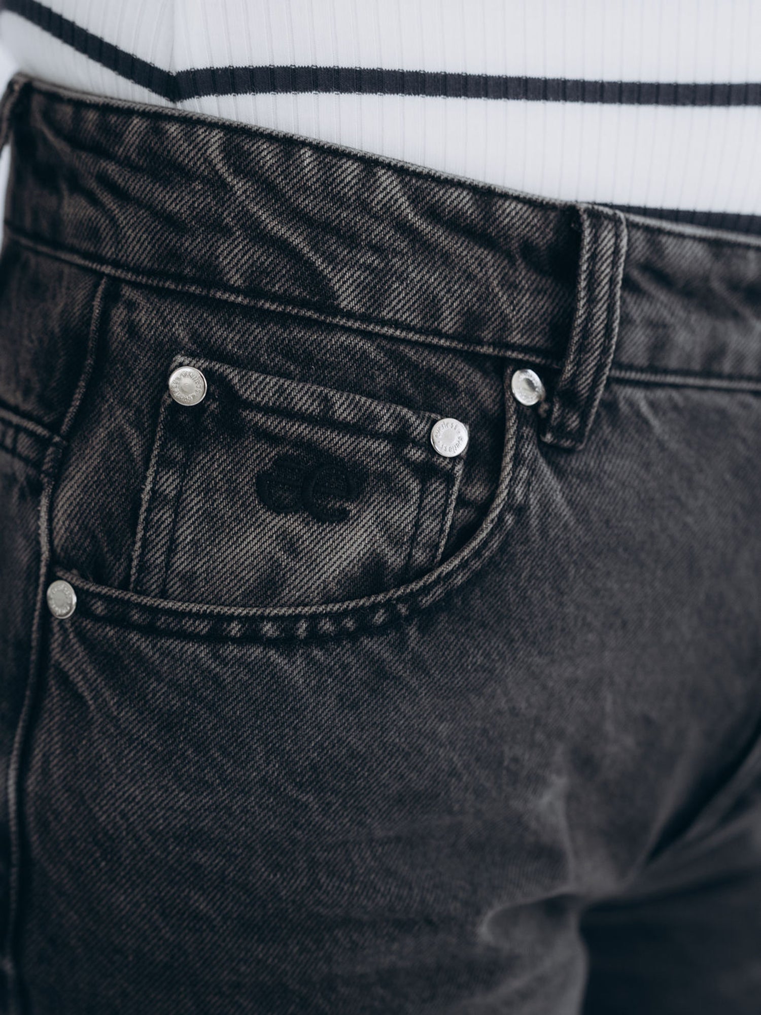 Detailaufnahme von Bund, Knopfverschluss und vorderen Taschen im verwaschenen dunkelgrauen Denim. Everless Signé eingestickt.