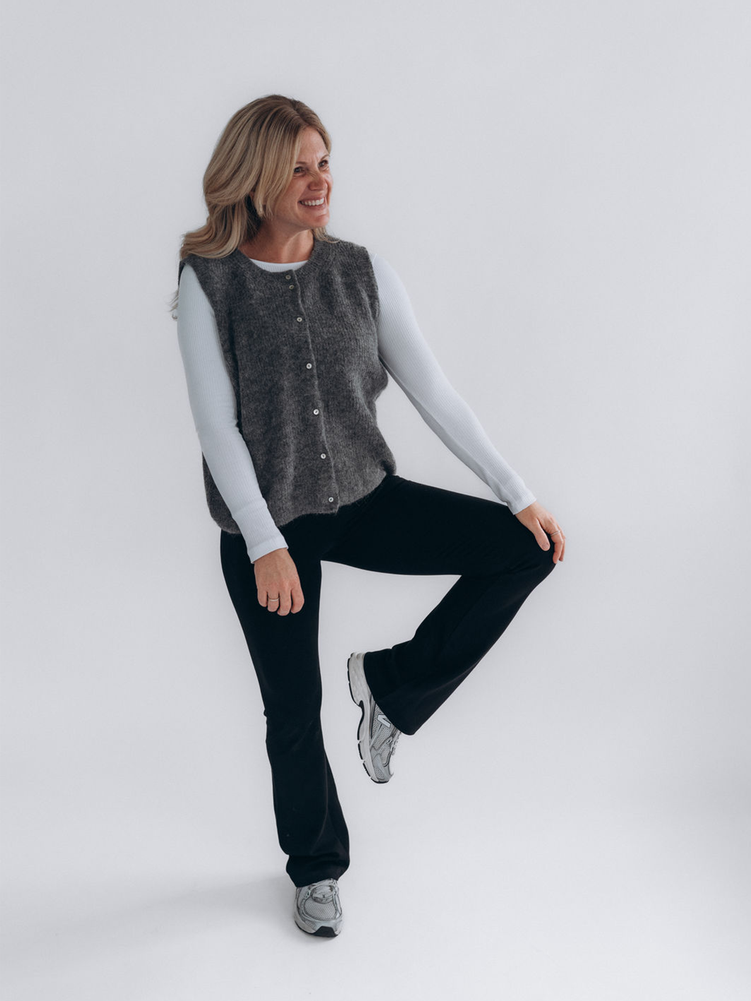 Angi trägt MARIE mit weißem Longsleeve LINNEA, grauer Strickweste LIV und Sneakern; ein Bein ist angewinkelt.