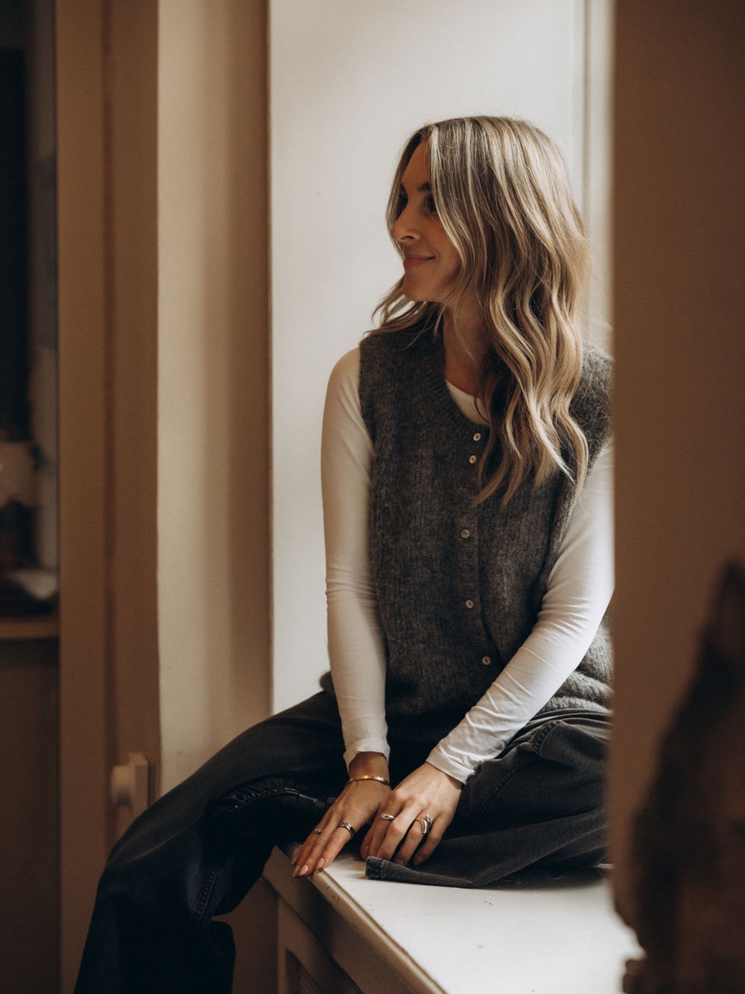 Lisa trägt die anthrazitfarbene Strickweste LIV über einem weißen Longsleeve, sitzend in einem warm beleuchteten Innenraum.
